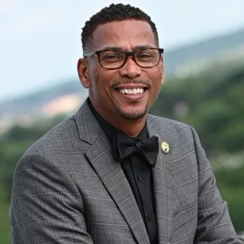 The image shows a smiling man wearing glasses, a black bow tie, and a gray suit. He appears to be in an outdoor setting with greenery in the background. The man has short, dark hair and a friendly expression. He seems to be posing for a portrait.